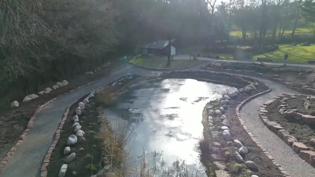 Luftaufnahme schräg: Teich mit Steinufer, Wege, Wiese und Bäume, im Hintergrund ein Holzhaus